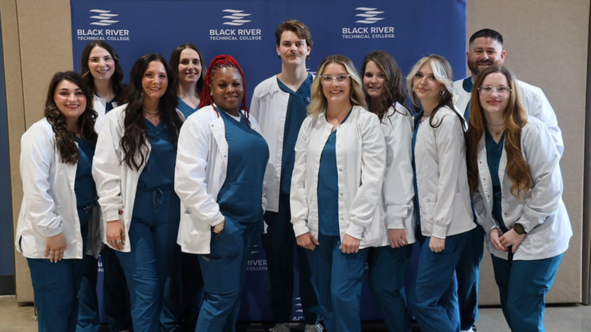 A group of nursing students in matching teal scrubs and white jackets pose together in front of a Black River Technical College backdrop.